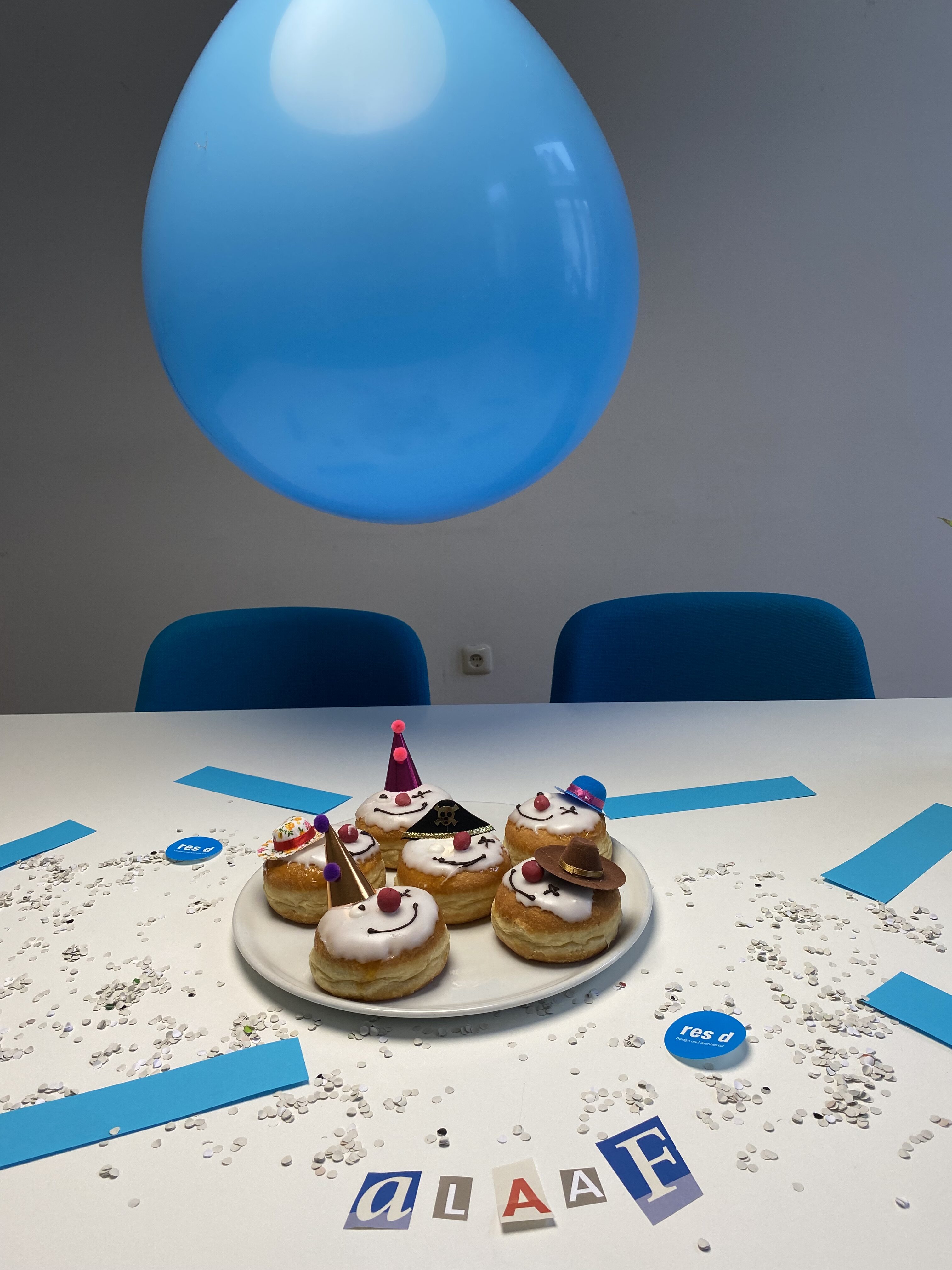 Muffins mit Clownsgesichtern darüber ein blauer Luftballon und rundherum Konfetti