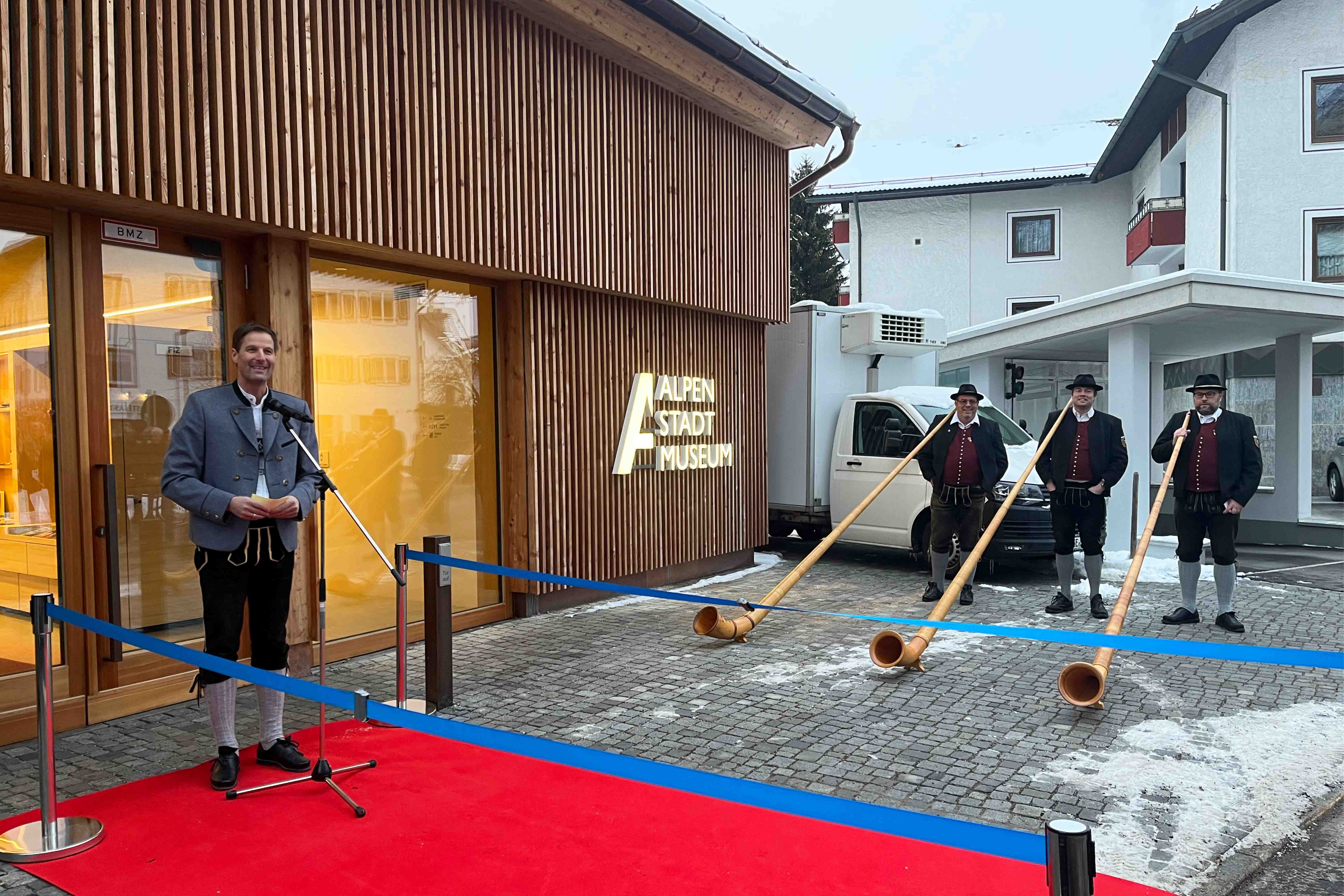 Eröffnungsrede des Bürgermeisters Christian Wilhelm vor dem Alpenstadtmuseum daneben stehen drei Männer mit jeweils einem Alphorn.